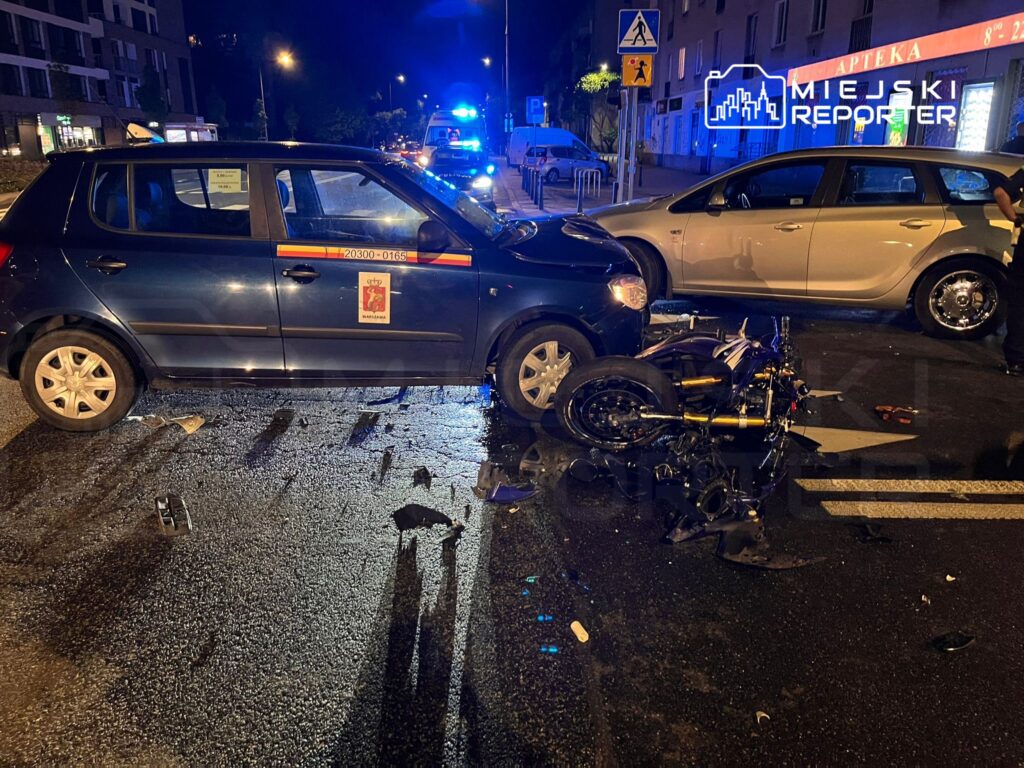 Czarny samochód osobowy z uszkodzeniami stoi na drodze obok przewróconego motocykla, w tle widać niebieskie światła policyjne.