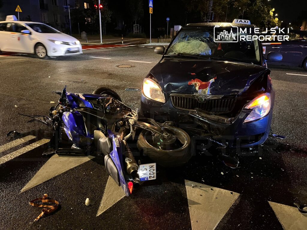 Motocykl leży na drodze obok zderzonego samochodu osobowego, w tle widać taksówkę i miejskie oświetlenie.