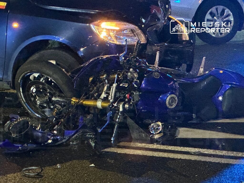 Motocykl w kolorze niebieskim leży na boku obok zderzaka samochodu, na mokrej nawierzchni drogi.