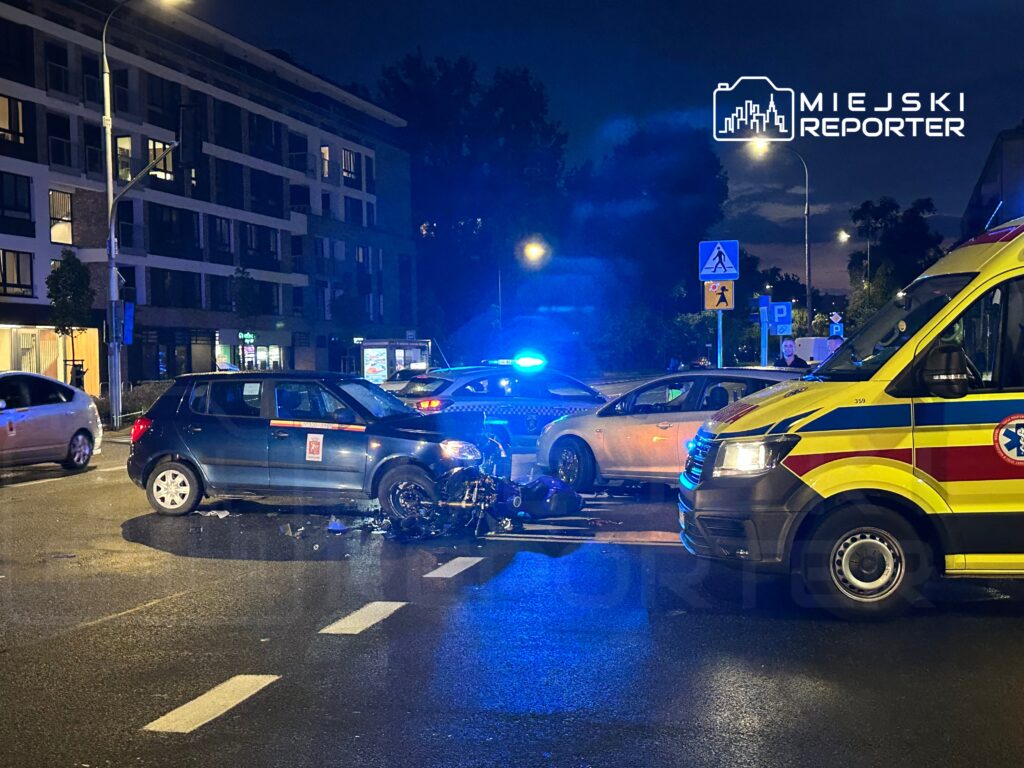 W nocy na ulicy stoi uszkodzony samochód osobowy, obok którego znajdują się pojazdy służb ratunkowych.