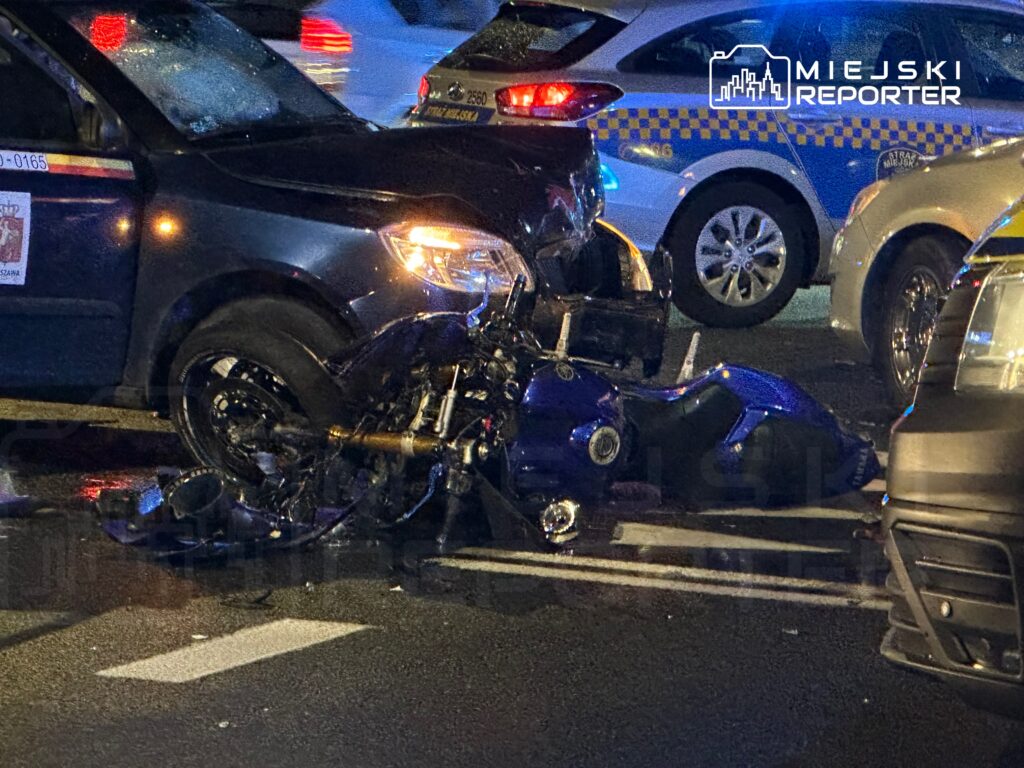 Motocykl leży na jezdni obok uszkodzonego samochodu osobowego, w tle widać inne pojazdy.