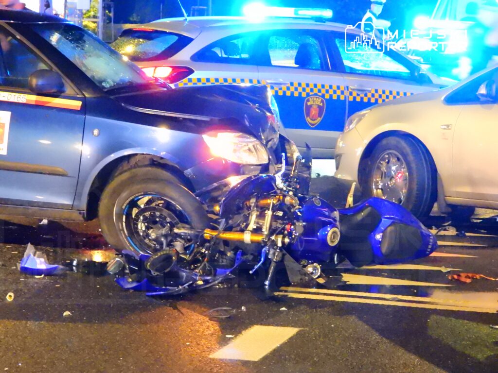 Motocykl leży na ziemi obok niebieskiego samochodu, w tle widoczne są nieoznakowane radiowozy policyjne.