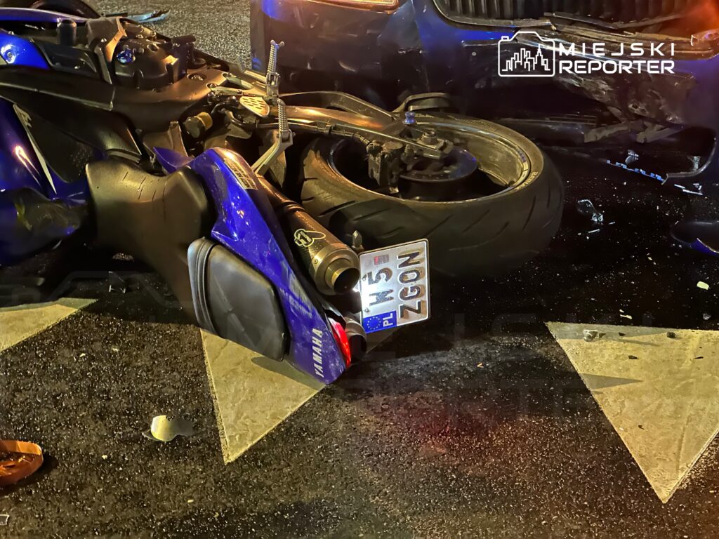 Motocykl leżący na boku na drodze, obok widoczny uszkodzony samochód. W tle widać asfalt i oznakowanie drogowe.
