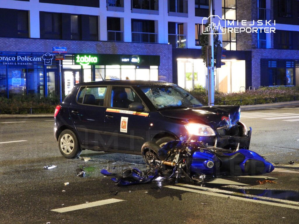 Samochód osobowy z uszkodzoną maską stoi na jezdni obok rozbitego motocykla, w tle widoczne oświetlone lokale.