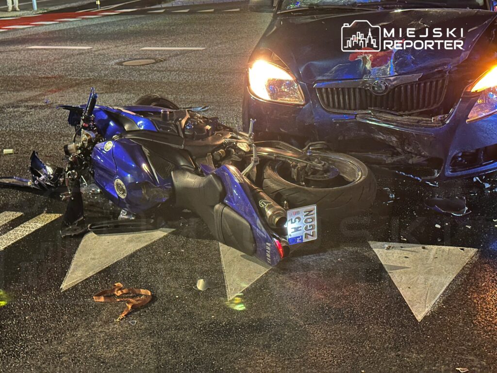 Motocykl leży na jezdni obok uszkodzonego samochodu osobowego po kolizji.