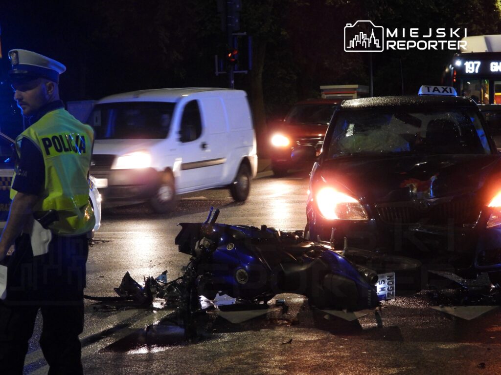 Policjant w żółtej kamizelce kieruje ruchem obok zderzonego motocykla i taksówki na mokrej jezdni.