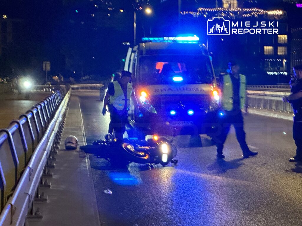 Na jezdni leży przewrócony motocykl, obok stoją ratownicy medyczni w kamizelkach odblaskowych, a w tle widać ambulans.