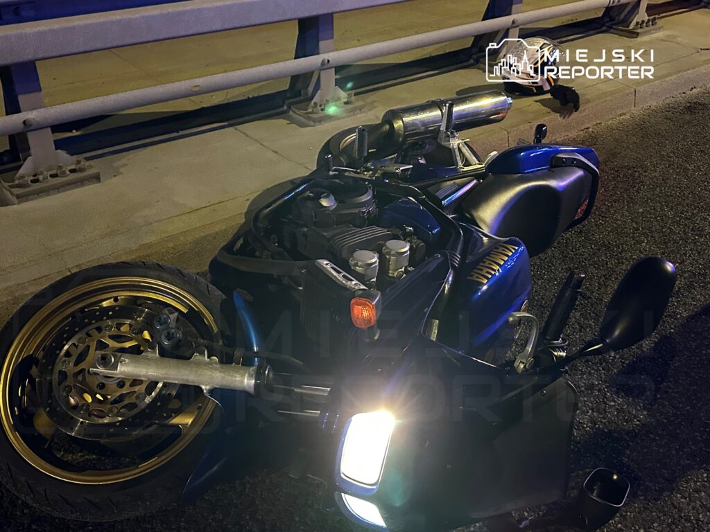 Motocykl leżący na boku na jezdni, z widocznymi uszkodzeniami oraz odblaskowym światłem.