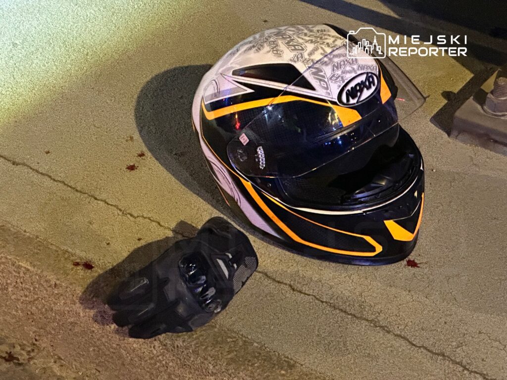 Kask motocyklowy z widocznymi śladami krwi leży na asfalcie obok rękawicy motocyklowej.
