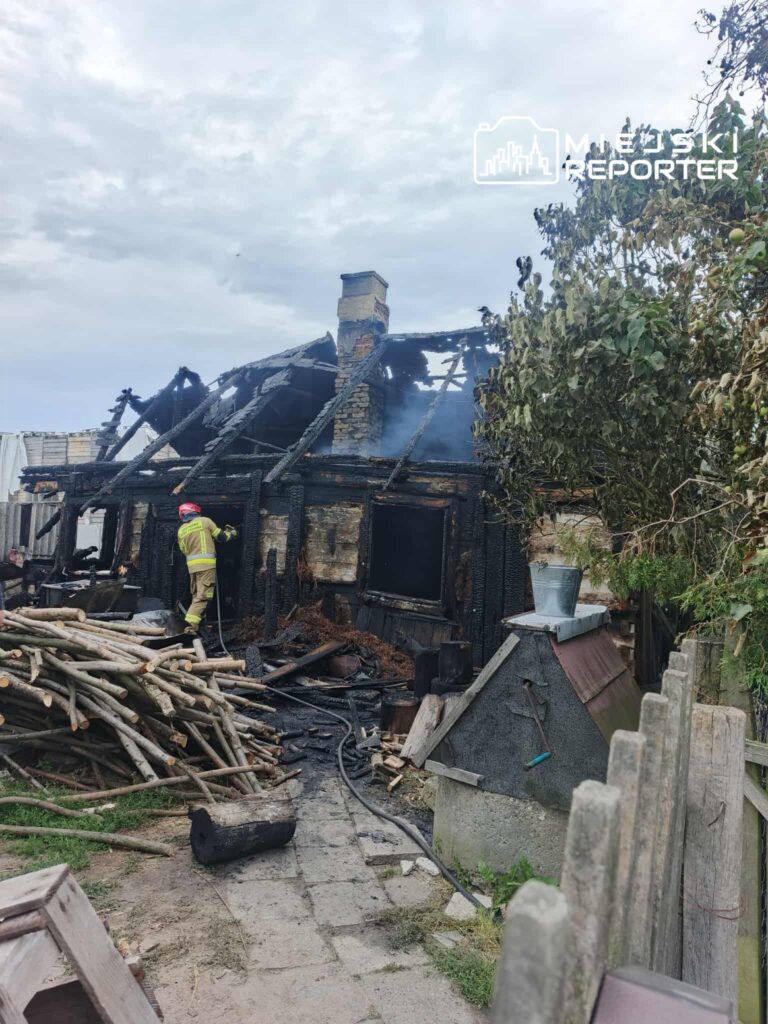 Strażak w żółtym mundurze przeszukuje zgliszcza spalonego domu, otoczonego stosami drewnianych bali i dymem.