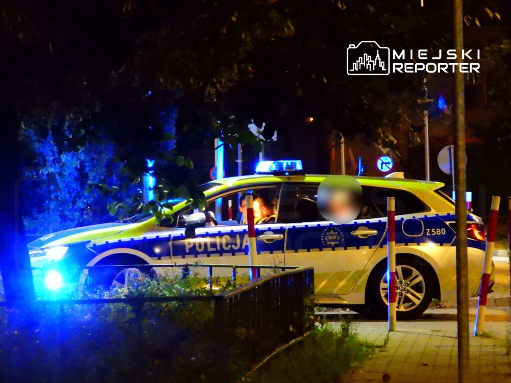 Policyjny radiowóz z włączonymi sygnałami świetlnymi stoi na ulicy nocą, a w środku znajdują się funkcjonariusze.