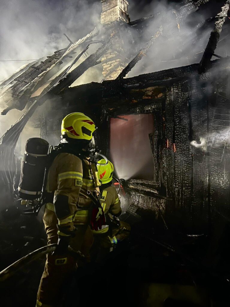 Dwaj strażacy w kombinezonach gaśniczych gaszą pożar w zniszczonym, zadymionym budynku.