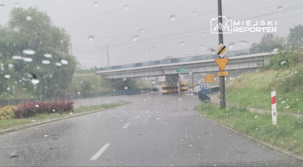 Intensywna ulewa powodująca podtopienie drogi w okolicy wiaduktu, widoczne kałuże i zamglony krajobraz.