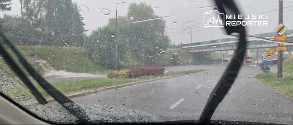 Intensywna ulewa z widocznymi kroplami deszczu na szybie samochodu, w tle zarośla i most.
