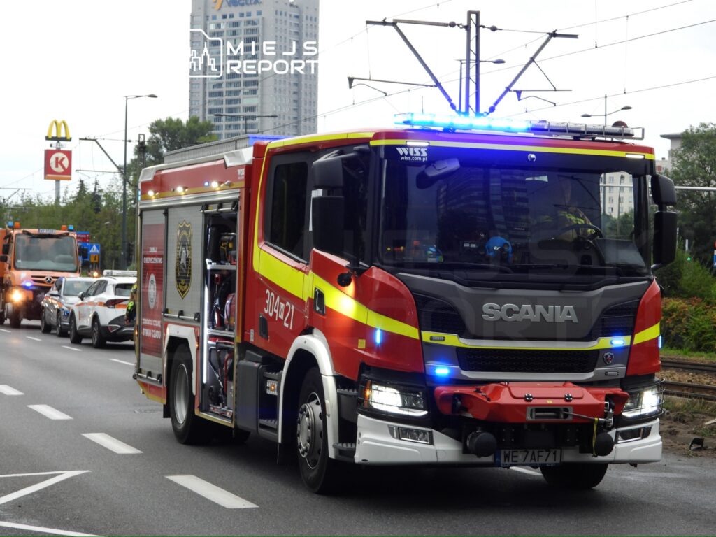 Czerwony wóz strażacki marki Scania z włączonymi sygnałami świetlnymi porusza się ulicą w miejskim otoczeniu.