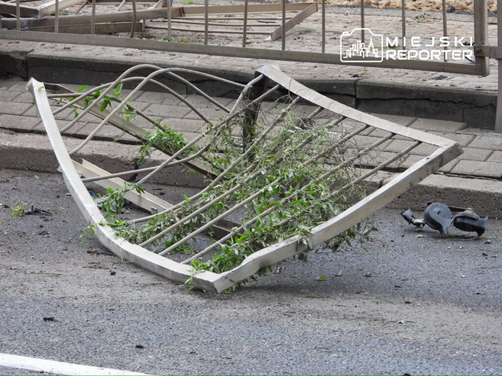 Złamana metalowa ławka z roślinnością leży na ulicy obok chodnika.
