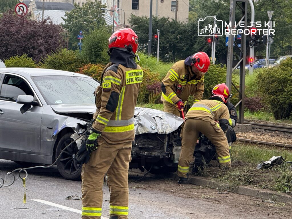Czterech strażaków w mundurach i hełmach pracuje przy rozbitym samochodzie na poboczu drogi, obok torów tramwajowych.