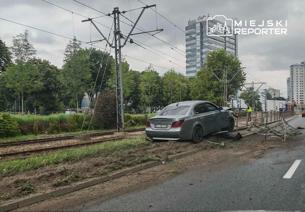 Samochód osobowy stoi na poboczu obok torów tramwajowych, w tle nowoczesne budynki i zieleń.