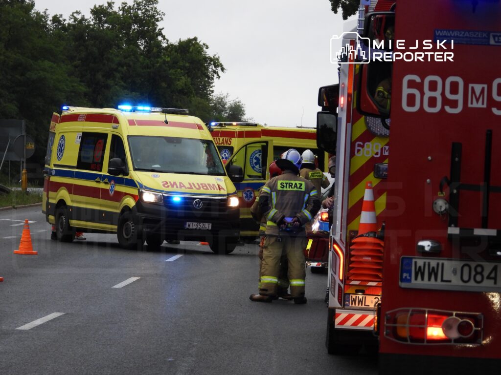 Zespół ratownictwa medycznego i strażacy współpracują podczas akcji ratunkowej na drodze, otoczeni przez pojazdy służb.