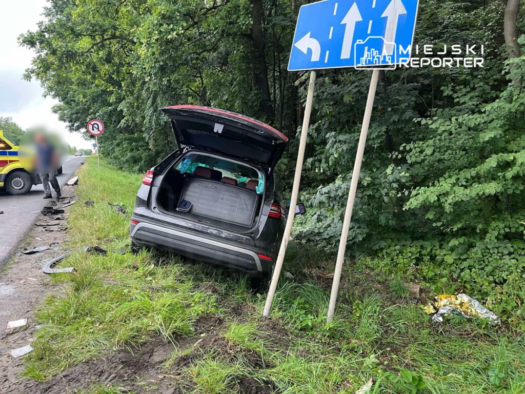 Czarny SUV zaparkowany na poboczu drogi z otwartą klapą bagażnika, obok znak drogowy z kierunkami.