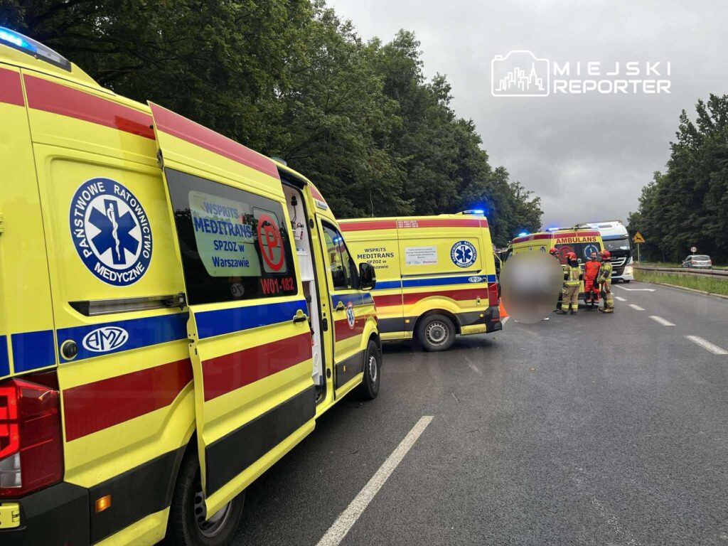 Zespół ratunkowy w żółtych ambulansach udziela pomocy na drodze, otoczony przez strażaków i sprzęt medyczny.