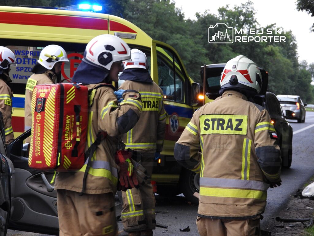 Grupa strażaków w mundurach z plecakami medycznymi ocenia sytuację przy wypadku drogowym obok karetki.