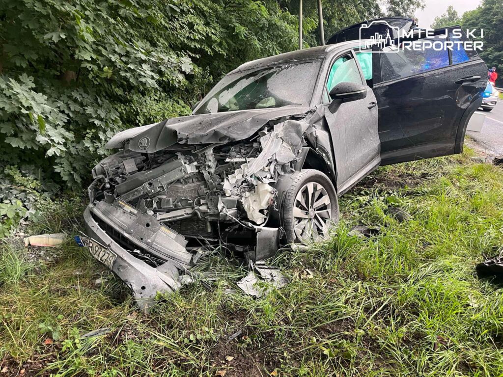 Zniszczony samochód osobowy stoi na poboczu drogi, obok gęstej roślinności.