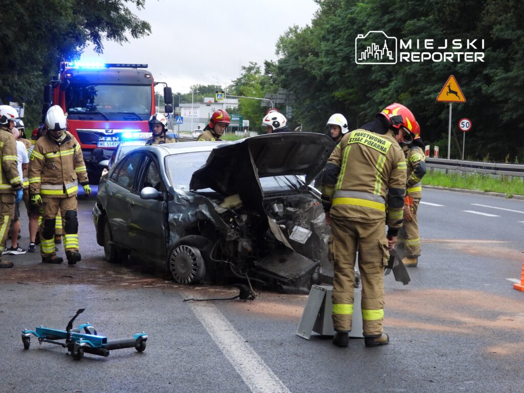 Grupa strażaków w mundurach pracuje przy uszkodzonym samochodzie na drodze, obok stoi wóz strażacki.