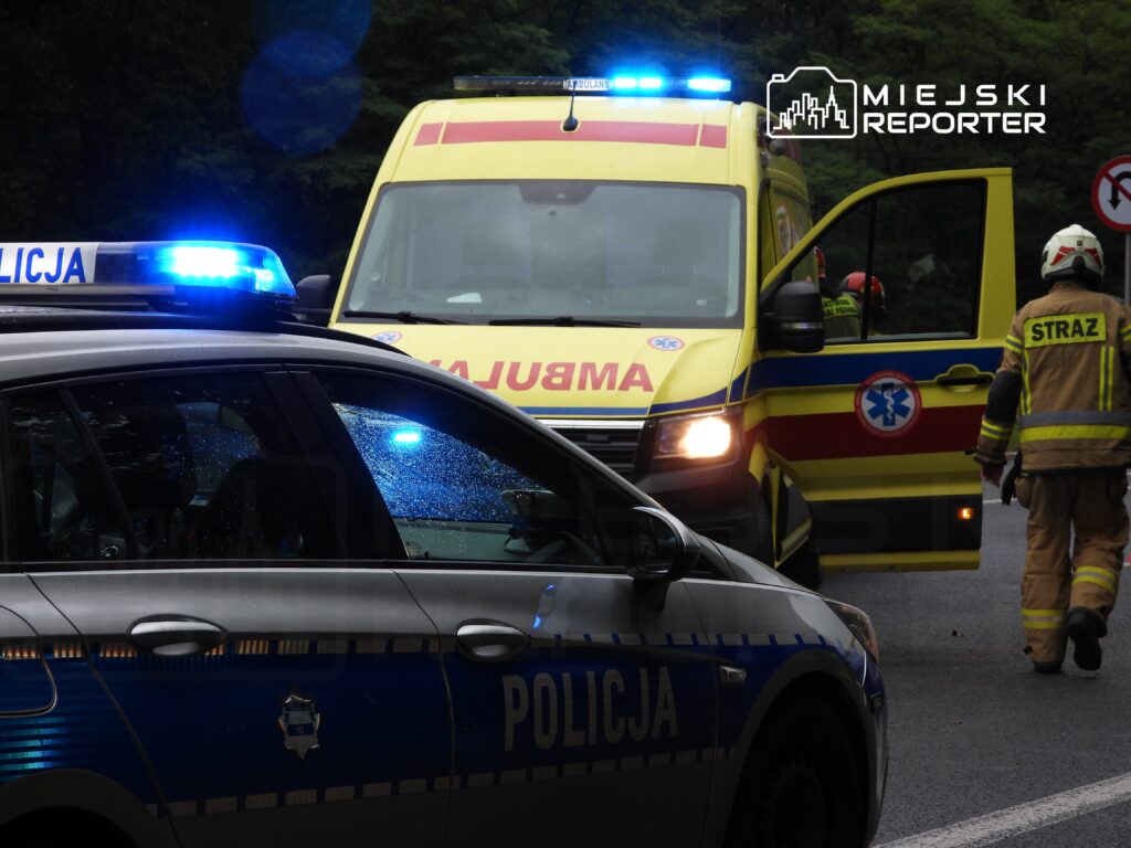 Radiowóz policyjny z włączonymi sygnałami świetlnymi stoi obok karetki pogotowia, a strażacy przystępują do akcji.