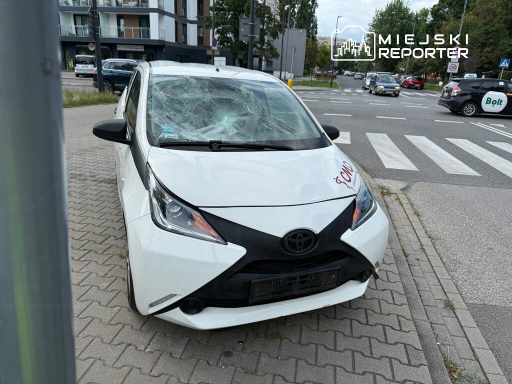 Uszkodzony biały samochód osobowy z rozbitą szybą przednią stoi na chodniku obok ruchliwej ulicy.
