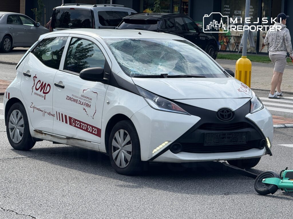 Uszkodzony samochód osobowy z wybitym przednim szybą stoi na ulicy, obok niego leży hulajnoga elektryczna.