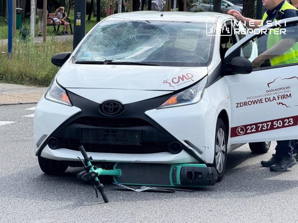 Zniszczony samochód osobowy z rozbitą szybą stoi na ulicy obok przewróconej hulajnogi elektrycznej, a obok policjant prowadzi czynności.