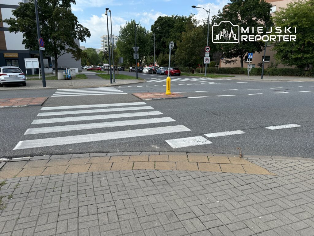 Pieszy przechodzi przez pasy w miejskim otoczeniu z widokiem na nowoczesne budynki i zaparkowane samochody.