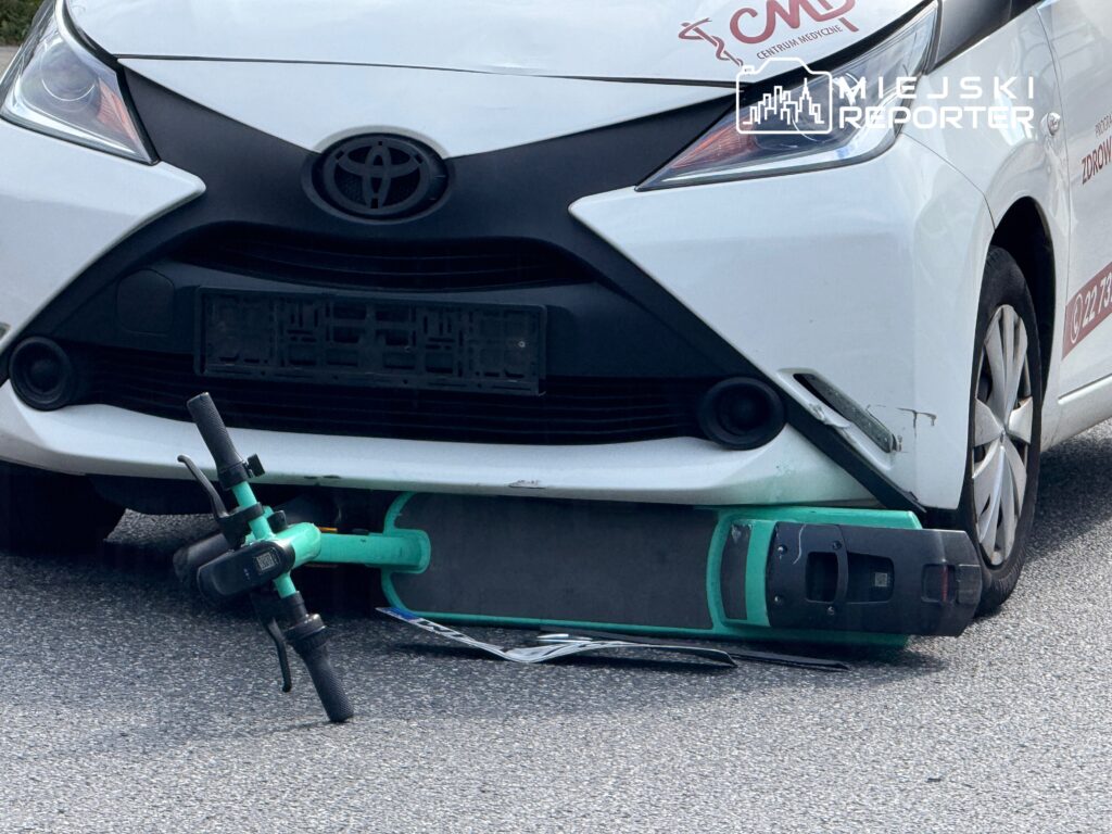 Zielona hulajnoga elektryczna leży pod przednim zderzakiem białego samochodu osobowego.