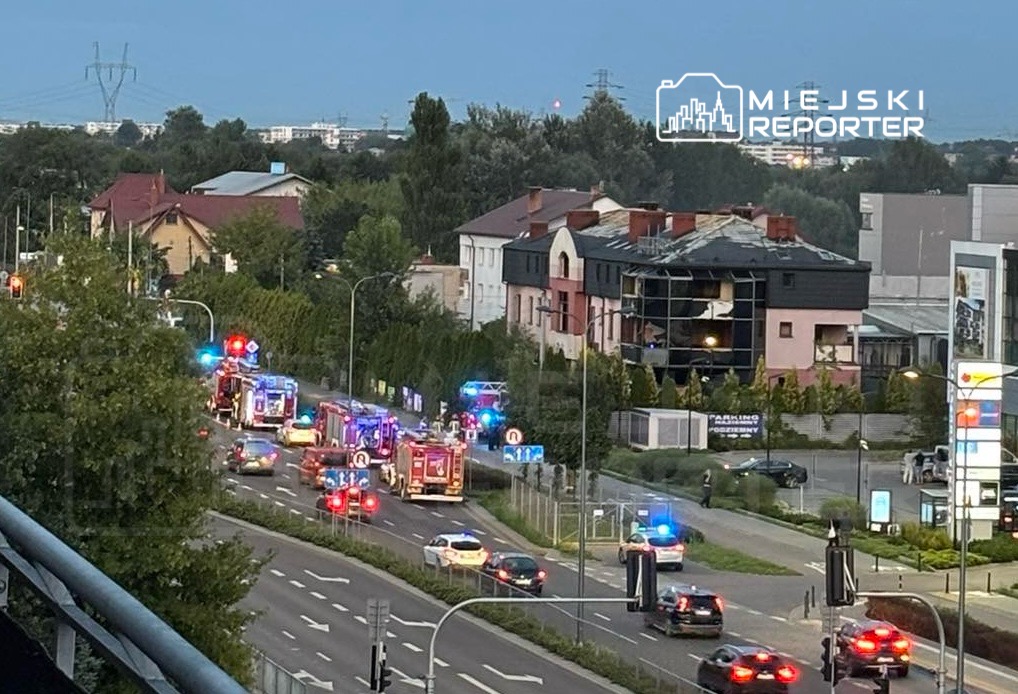 Kilka wozów strażackich z włączonymi sygnałami świetlnymi na ruchliwej ulicy obok uszkodzonego budynku.