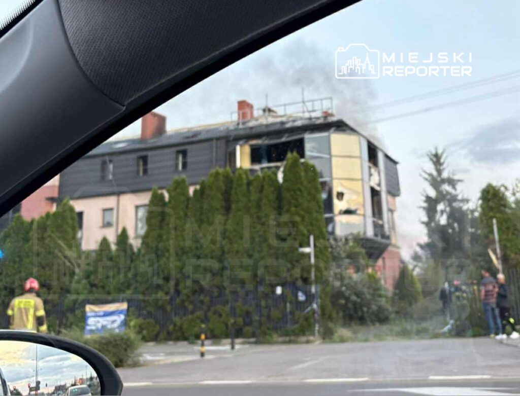 Pożar w budynku mieszkalnym, z którego wydobywa się dym, w otoczeniu drzew i osób obserwujących zdarzenie.