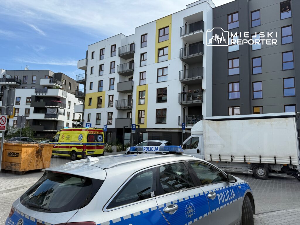 Policyjny radiowóz i ambulans stoją na osiedlu mieszkaniowym z nowoczesnymi budynkami w tle.