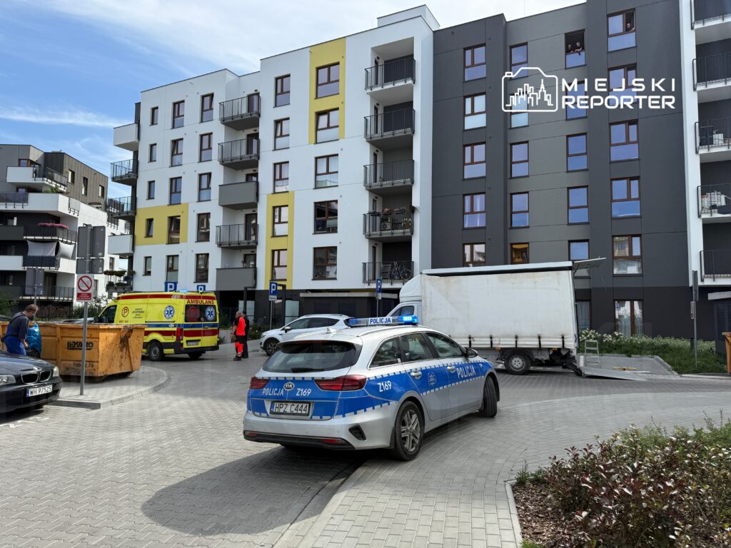 Karetka pogotowia i radiowóz policji stoją na podjeździe nowoczesnego budynku mieszkalnego, obok kontenera na odpady.