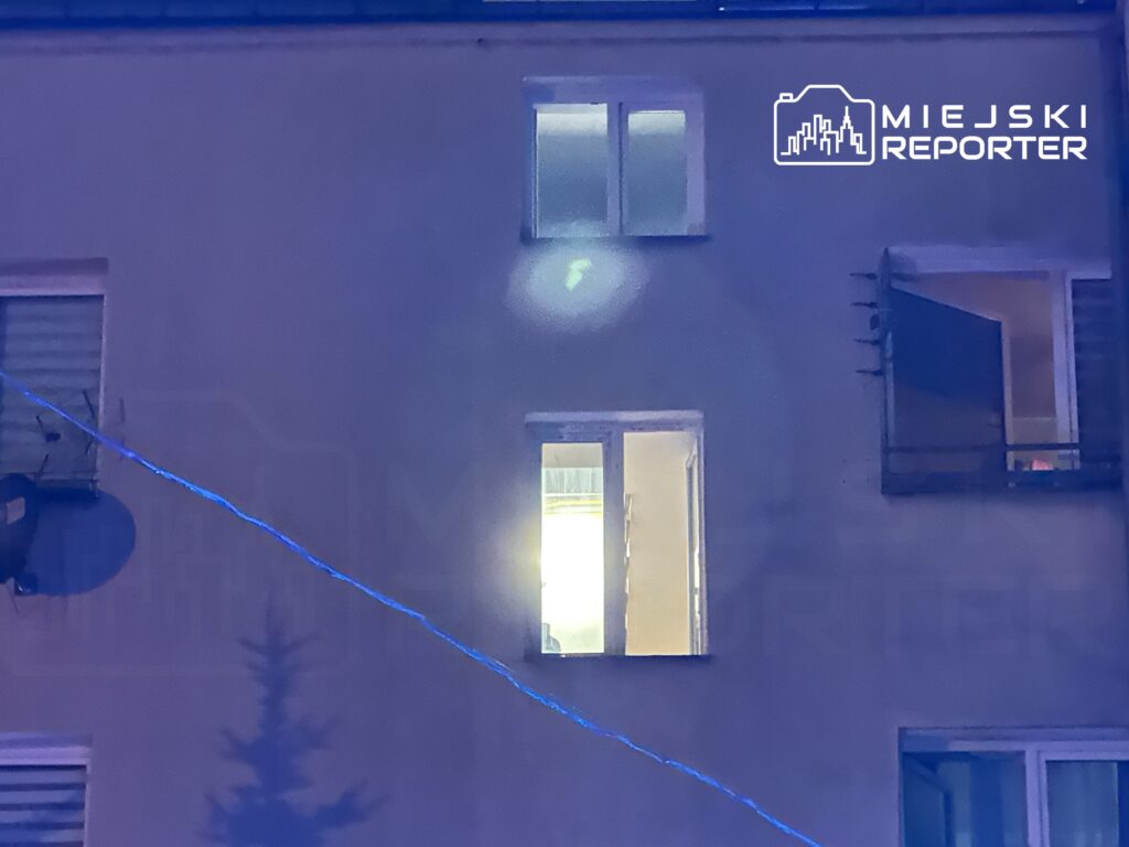 Oświetlone okno mieszkania na tle ciemnej elewacji budynku, z widocznym przewodem elektrycznym.