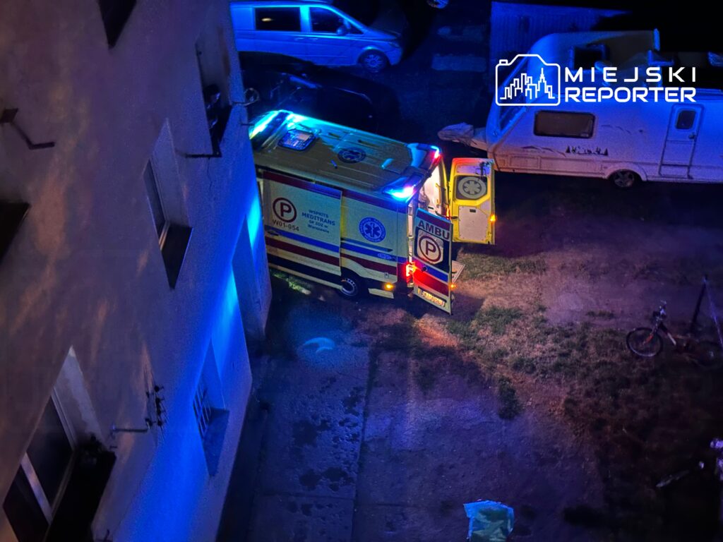 Karetka pogotowia z włączonymi sygnałami świetlnymi stoi na podjeździe obok budynku mieszkalnego.