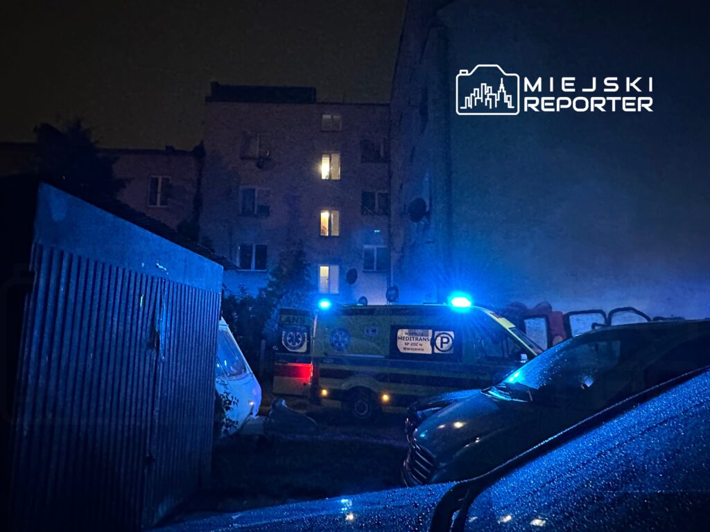 Nocna scena z niebieskimi światłami karetki w wąskiej uliczce, otoczonej budynkami i zaparkowanymi samochodami.