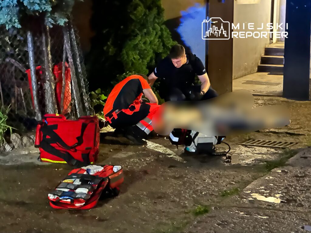 Ratownicy medyczni udzielają pomocy poszkodowanemu na chodniku, obok torów i ciemnej ściany budynku.