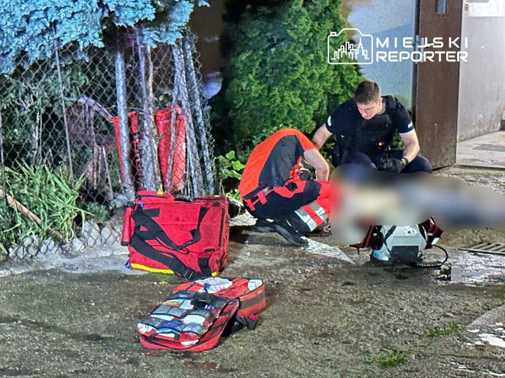 Ratownicy medyczni udzielają pomocy poszkodowanemu na chodniku, obok czerwonych torb ratunkowych.