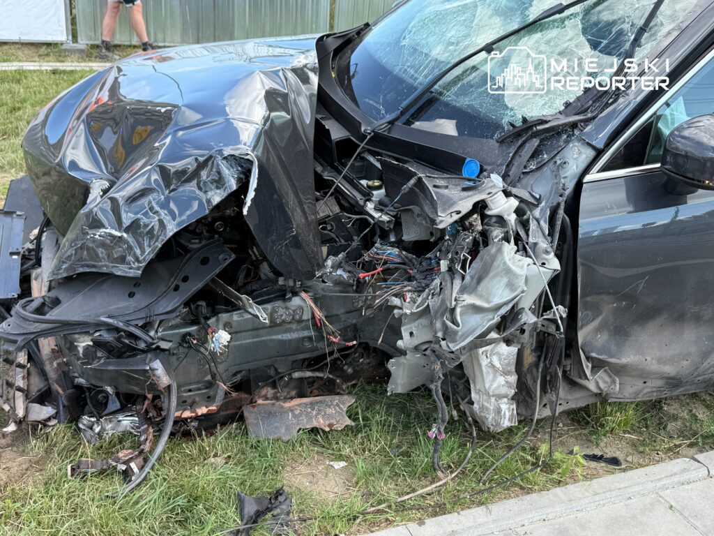 Zniszczony przód samochodu po wypadku, z widocznymi uszkodzeniami blacharskimi i odsłoniętymi przewodami, na tle trawnika.