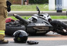Motocykl marki Yamaha leży na boku na jezdni, obok niego znajduje się kask z logo Monster. W tle widoczny strażak w mundurze.