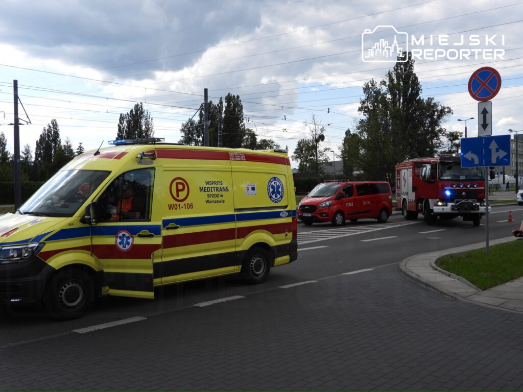 Karetka pogotowia z włączonymi sygnałami przejeżdża obok straży pożarnej i samochodu dostawczego na skrzyżowaniu.