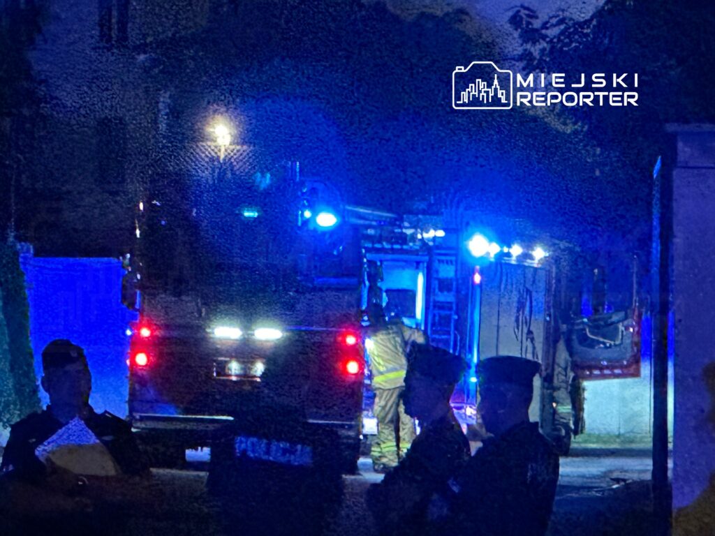 Strażacy w mundurach wchodzą do wozu strażackiego, a policjanci obserwują sytuację w nocy.