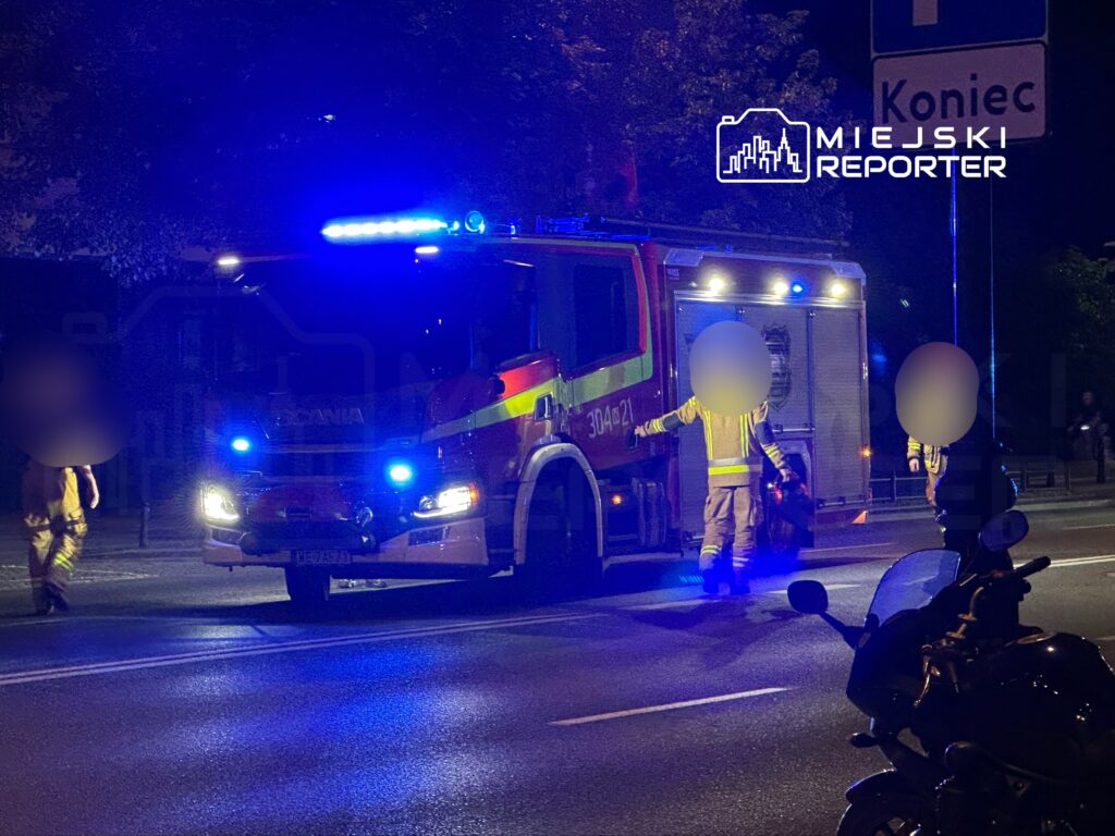 Strażacy w mundurach zbliżają się do wozu strażackiego na oświetlonej ulicy nocą.