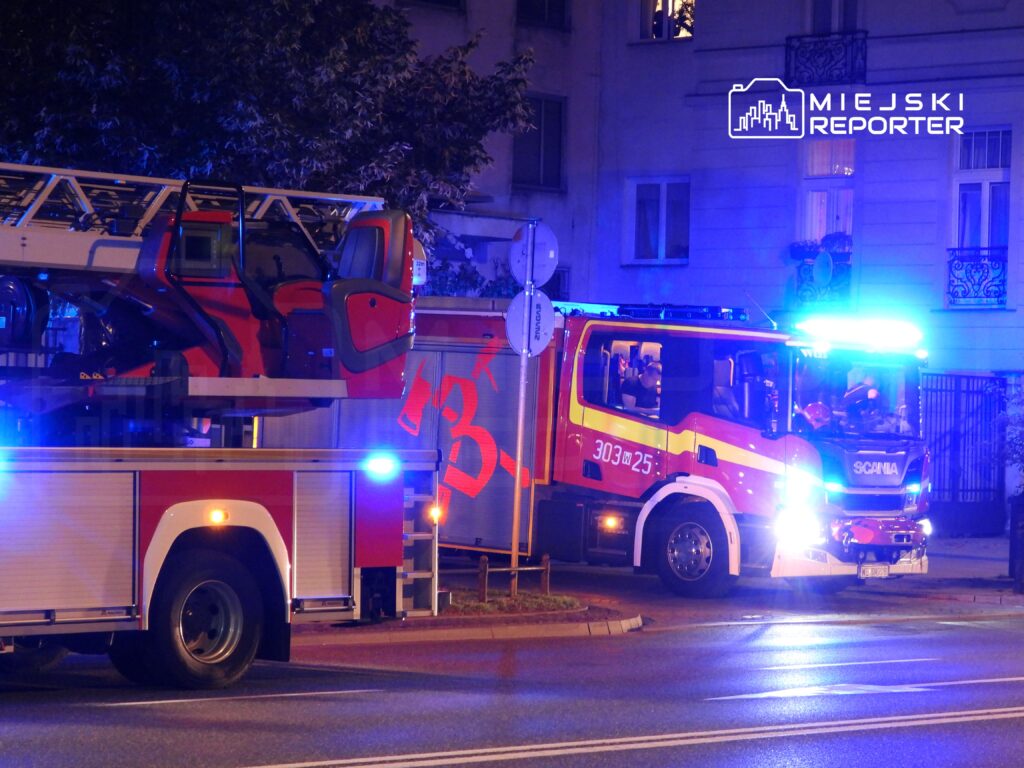 Straż pożarna z włączonymi sygnałami świetlnymi stoi na ulicy, obok wozu strażackiego z podnośnikiem.