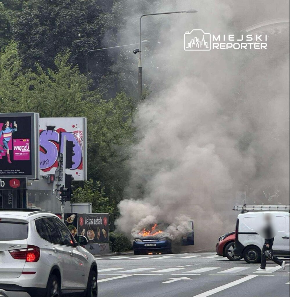Samochód zajęty ogniem na ulicy, w tle gęsty dym i billboardy reklamowe.